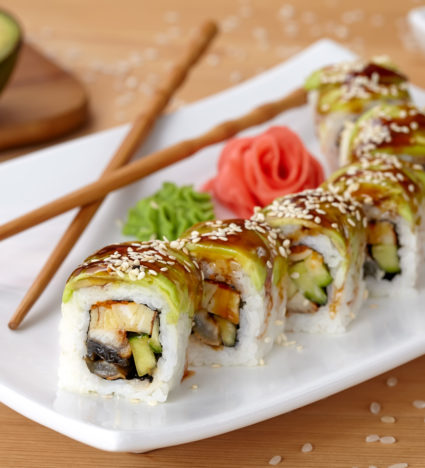 Green dragon sushi roll with eel, avocado, cucumber, wasabi and ginger. Traditional asian rice sushi healthy seafood. White plate, wooden table background.
